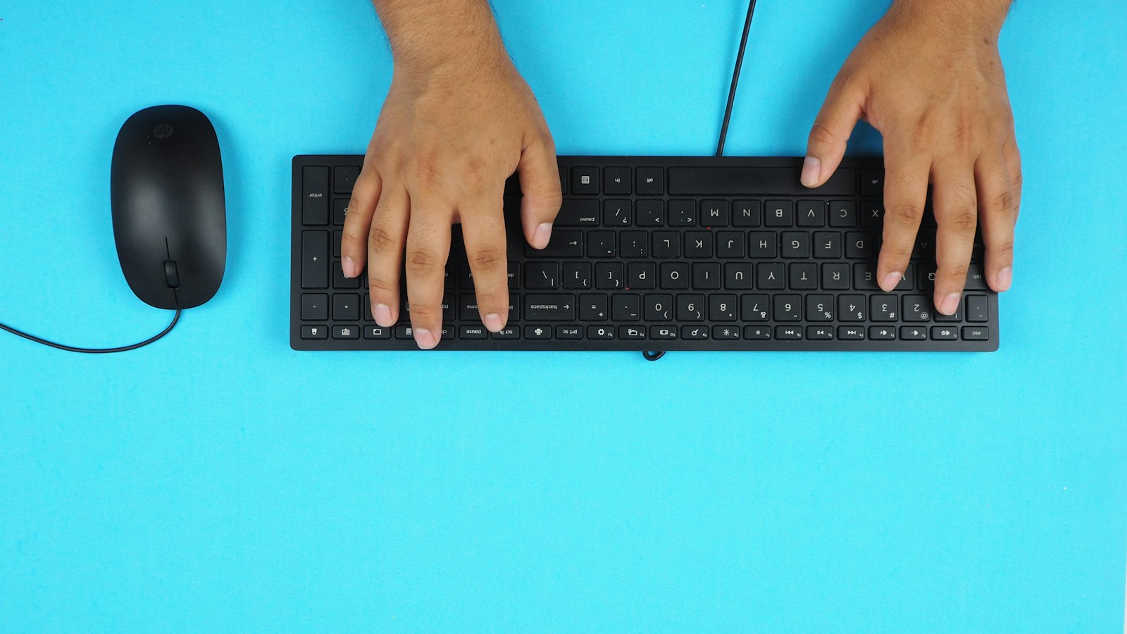 Hands typing on a black keyboard next to a mouse on a blue desk (photo: Luis Villafranca / Unsplash)