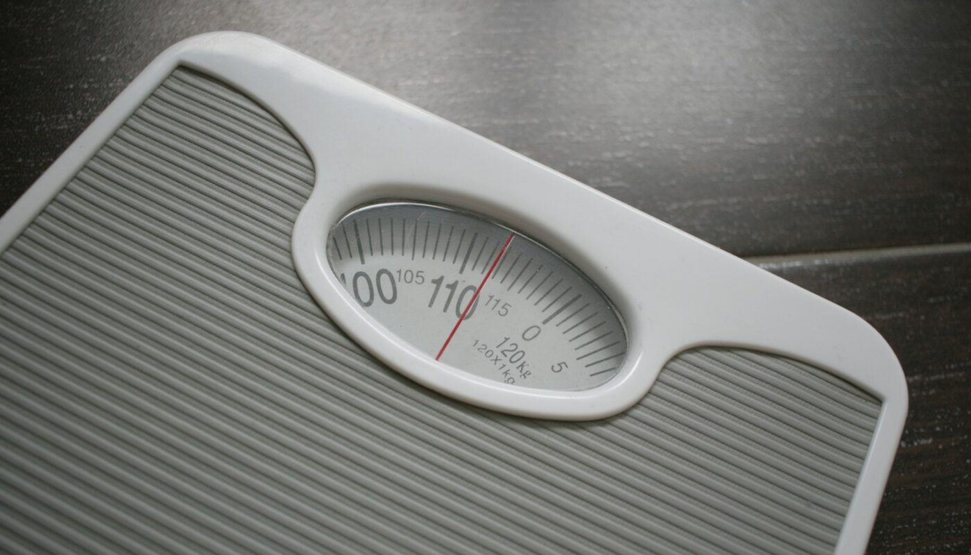 A close view of a white analogue bathroom scale with a red needle and printed dial (photo: Joachim Schnurle / Unsplash)