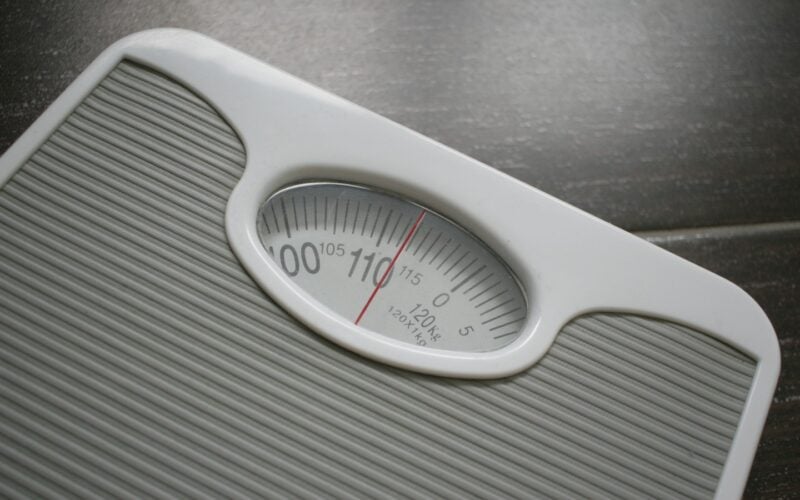 A close view of a white analogue bathroom scale with a red needle and printed dial (photo: Joachim Schnurle / Unsplash)