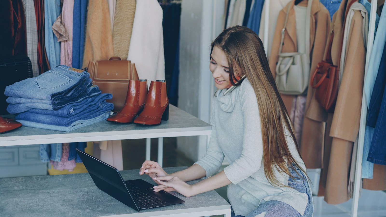 A boutique owner smiling on the phone while working on a laptop among rails of clothes, folded jeans and boots (photo: Vitaly Gariev / Unsplash)