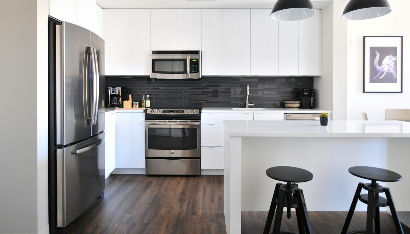 A bright white kitchen with a stainless steel fridge, range and over-the-range microwave, and two black stools at the island (photo: Naomi Hébert / Unsplash)