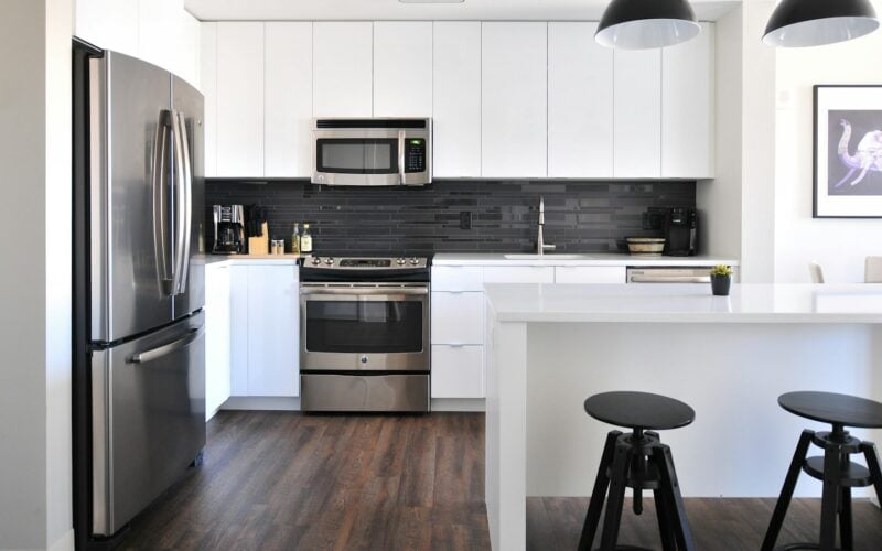 A bright white kitchen with a stainless steel fridge, range and over-the-range microwave, and two black stools at the island (photo: Naomi Hébert / Unsplash)