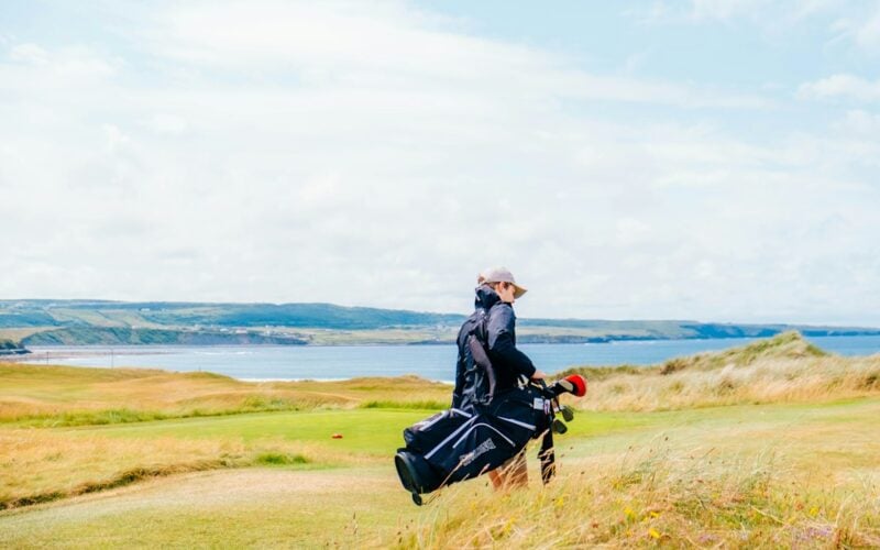 A golfer carries a stand bag on their shoulder while walking a windswept coastal links fairway above the sea.