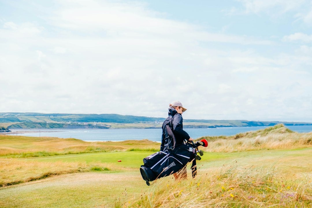A golfer carries a stand bag on their shoulder while walking a windswept coastal links fairway above the sea.