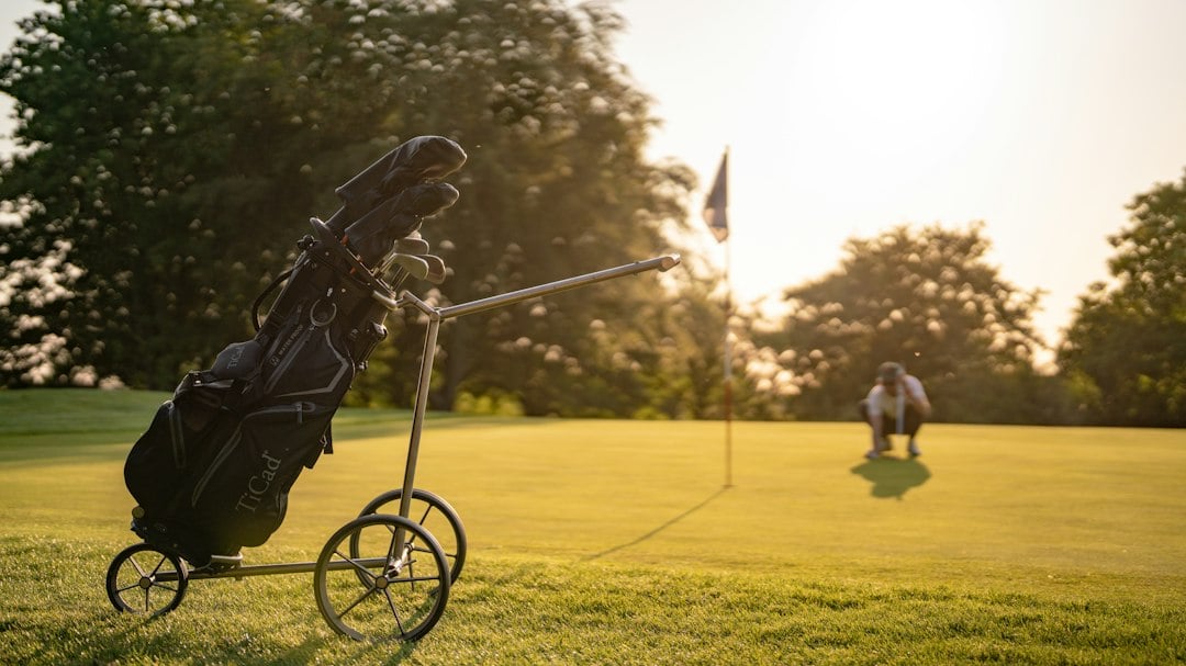 A golf bag sits on a push trolley beside a green at sunset while a player tends the flagstick in the distance.