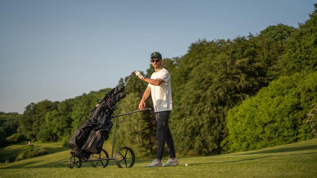 A golfer stands on the tee next to a three-wheel push trolley holding his golf bag, with trees in the background.