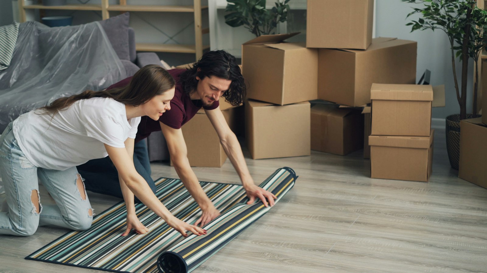 A couple kneeling to unroll a striped rug in a living room stacked with moving boxes (photo: Vitaly Gariev / Unsplash)