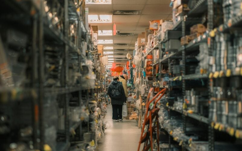 A lone shopper walking down a narrow store aisle lined floor to ceiling with stock (photo: Tianlei Wu / Unsplash)