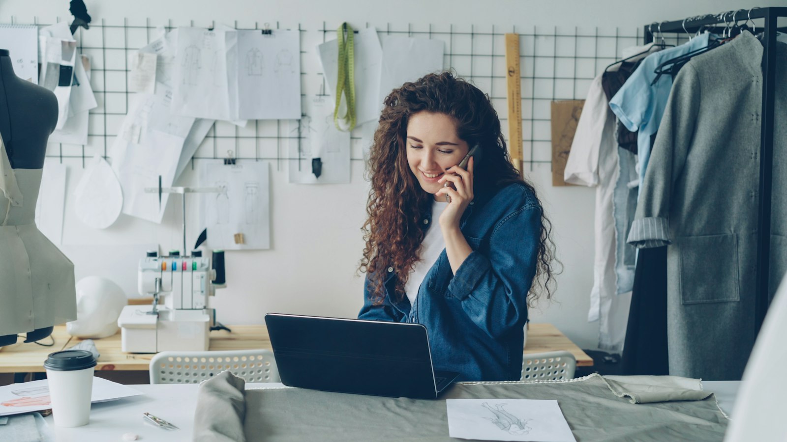 A fashion designer smiling on the phone while working at a laptop in her studio, with sketches pinned to the wall behind her (photo: Vitaly Gariev / Unsplash)