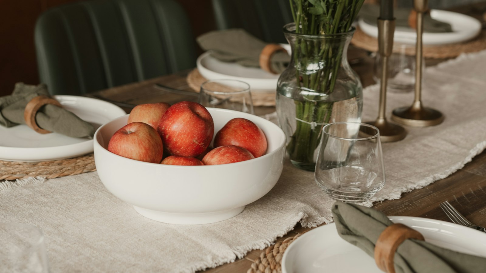 A dining table laid with white plates, linen napkins, a bowl of red apples and a vase of red flowers (photo: Clay Banks / Unsplash)
