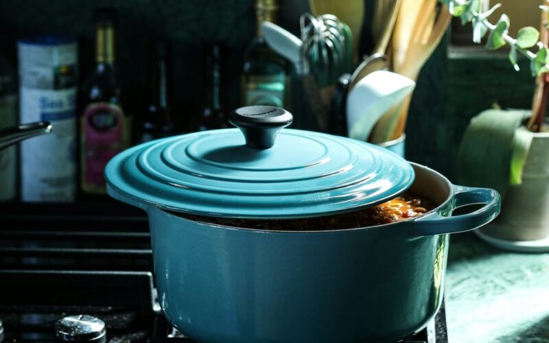 Teal enamelled cast-iron Dutch oven simmering on a gas hob in a dark green kitchen