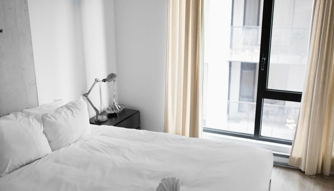 An empty white hotel bed beside a window (photo: Andrew Neel / Unsplash)