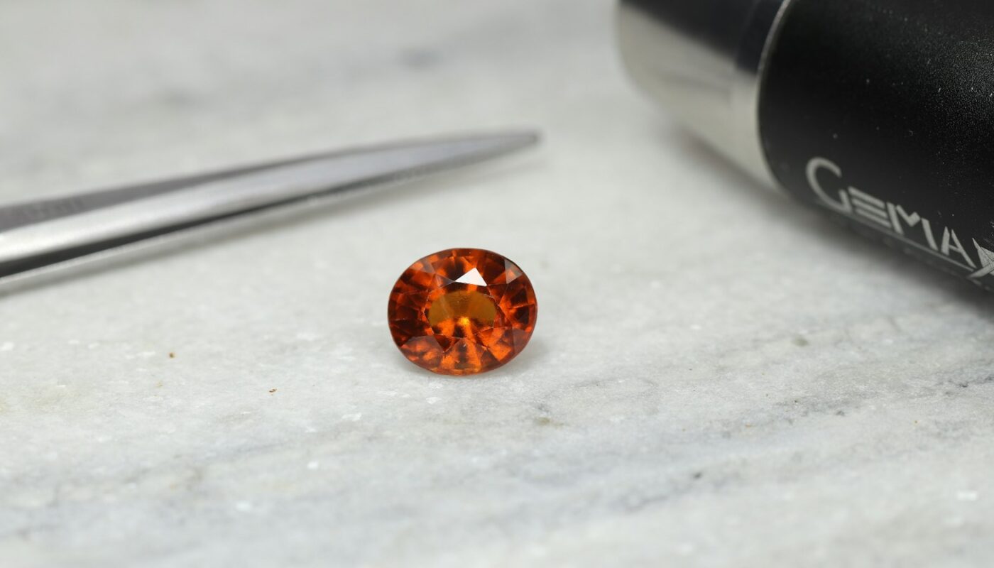 An orange faceted gemstone resting on a grey surface next to jeweller’s tweezers and a gem-testing instrument (photo: sameer Ali / Unsplash)