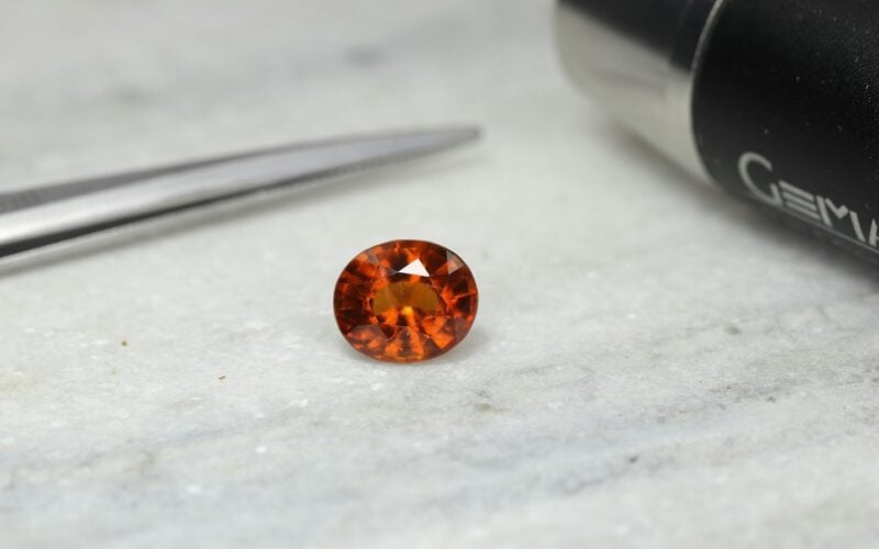 An orange faceted gemstone resting on a grey surface next to jeweller’s tweezers and a gem-testing instrument (photo: sameer Ali / Unsplash)