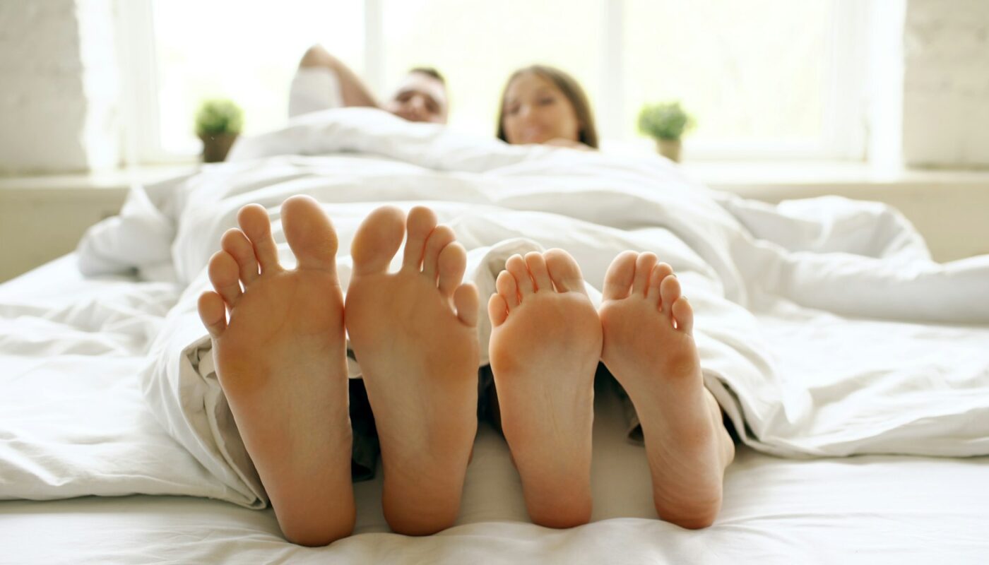 Two pairs of feet poking out from beneath a white duvet at the end of a bed (photo: Vitaly Gariev / Unsplash)