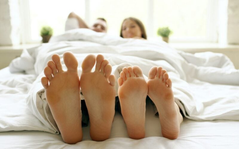 Two pairs of feet poking out from beneath a white duvet at the end of a bed (photo: Vitaly Gariev / Unsplash)
