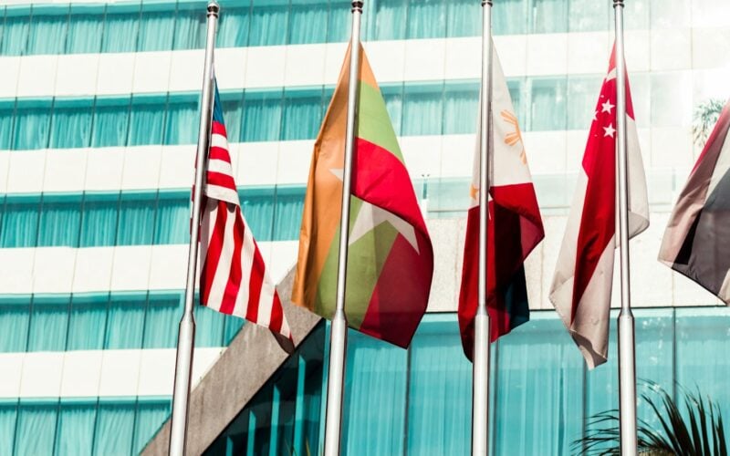Flags of several countries flying in front of a glass office building (photo: Kaung Myat Min / Unsplash)