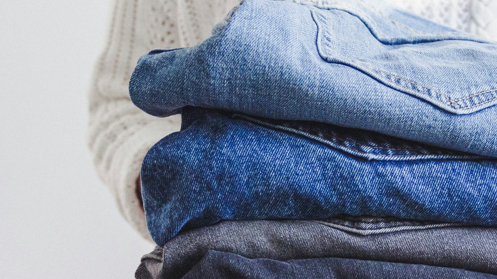 A person in a cream cable-knit sweater holding a pile of folded jeans in different washes (photo: Maude Frédérique Lavoie / Unsplash)