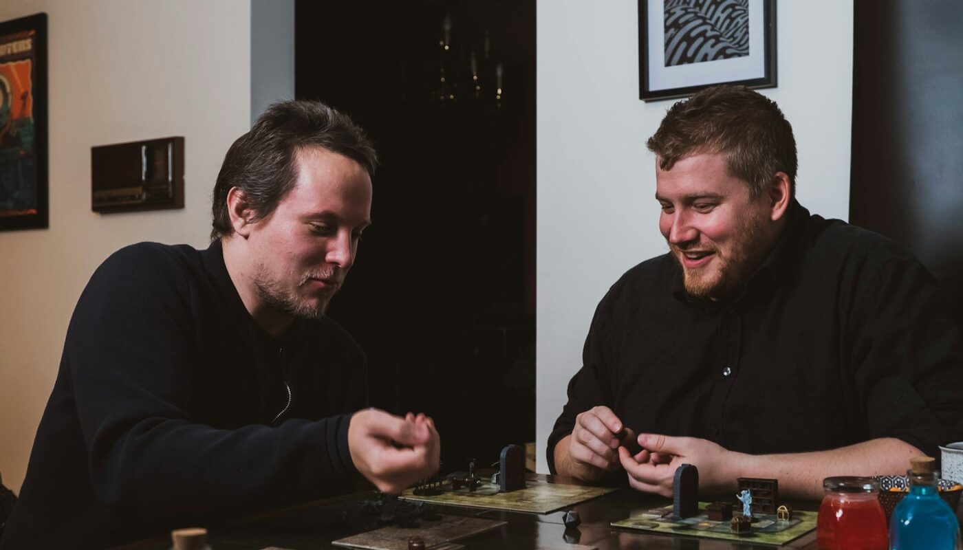 Two friends playing a miniatures board game with dice at a wooden table (photo: 2H Media / Unsplash)