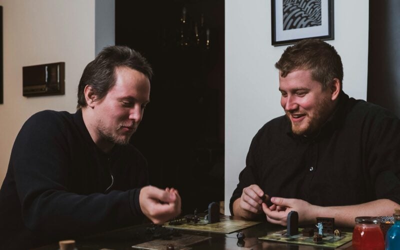 Two friends playing a miniatures board game with dice at a wooden table (photo: 2H Media / Unsplash)