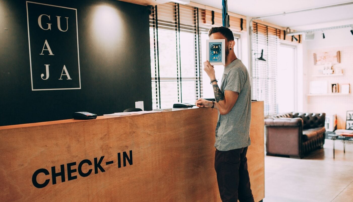 A guest standing at a hotel check-in desk holding a phone (photo: Helena Lopes / Unsplash)