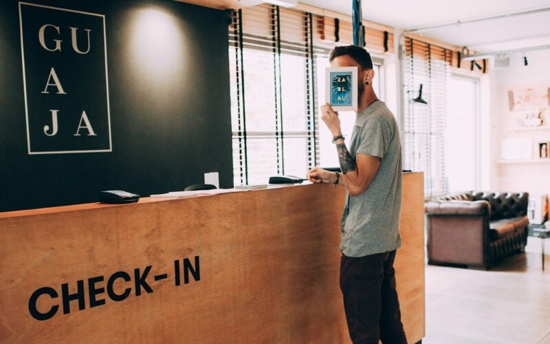A guest standing at a hotel check-in desk holding a phone (photo: Helena Lopes / Unsplash)