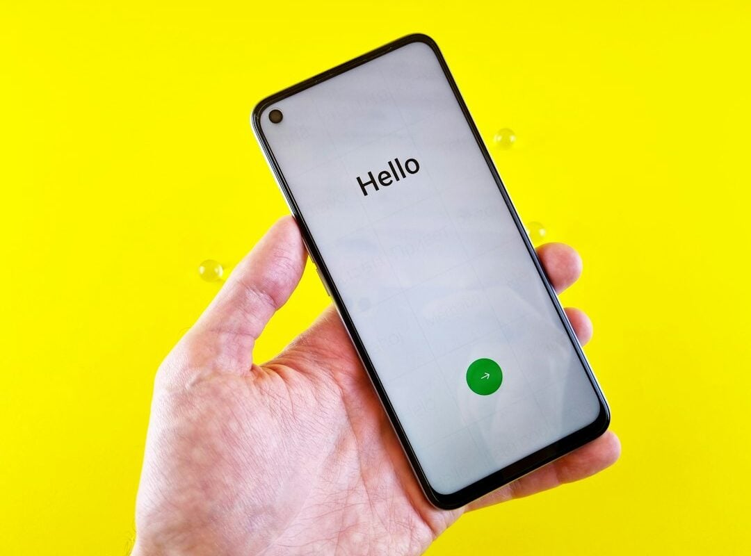 A hand holds a smartphone displaying the word Hello on its setup screen against a yellow backdrop