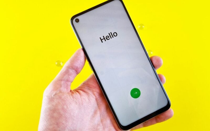 A hand holds a smartphone displaying the word Hello on its setup screen against a yellow backdrop