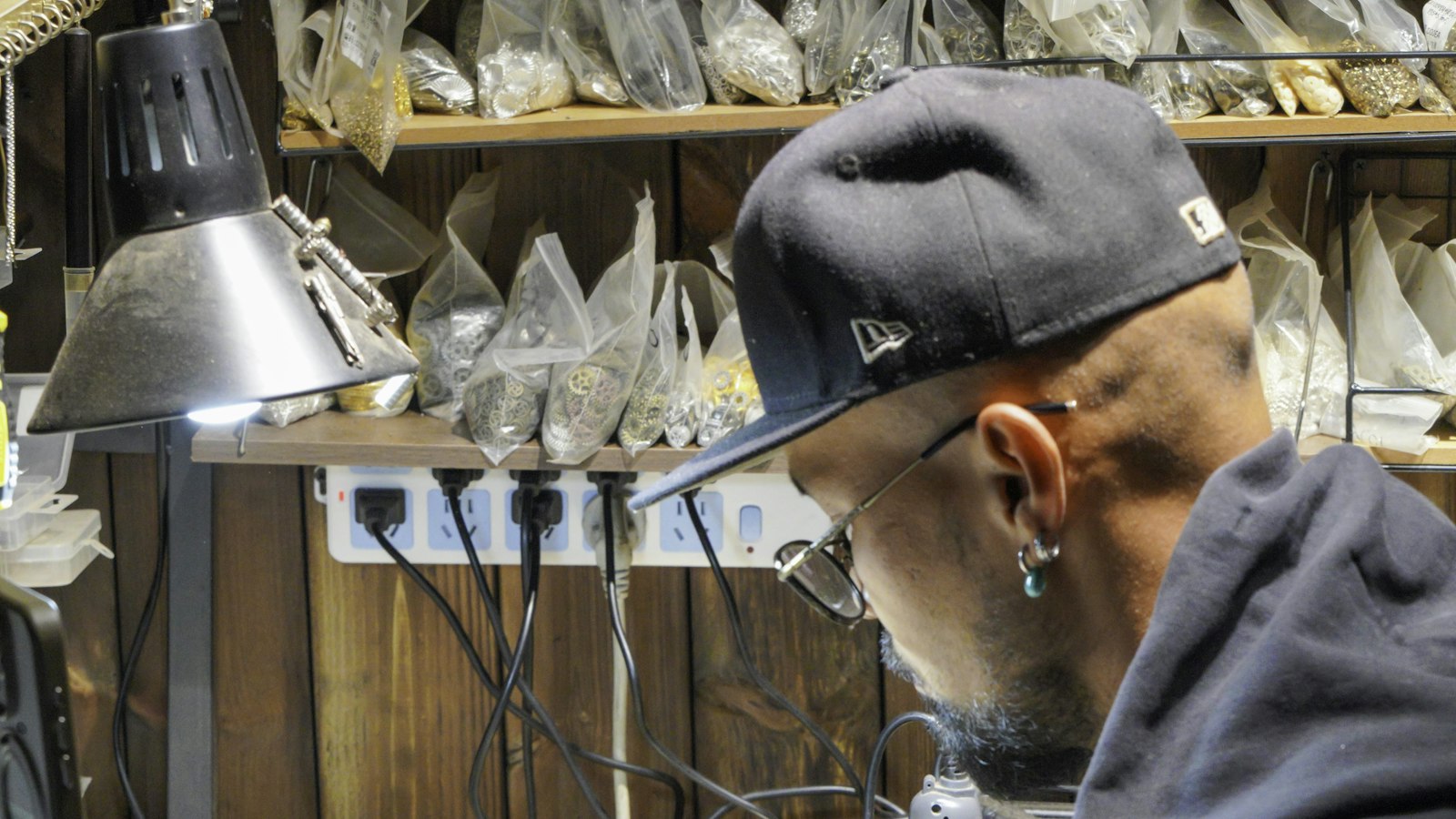 A jeweller in a cap working on small pieces at a crowded workbench strewn with tools and trays