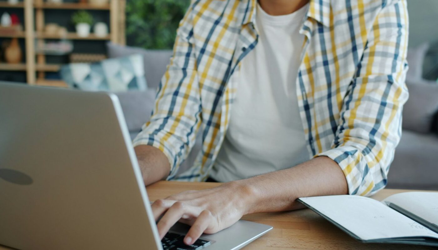 Man typing on a laptop beside an open notebook at a desk (photo: Vitaly Gariev / Unsplash)