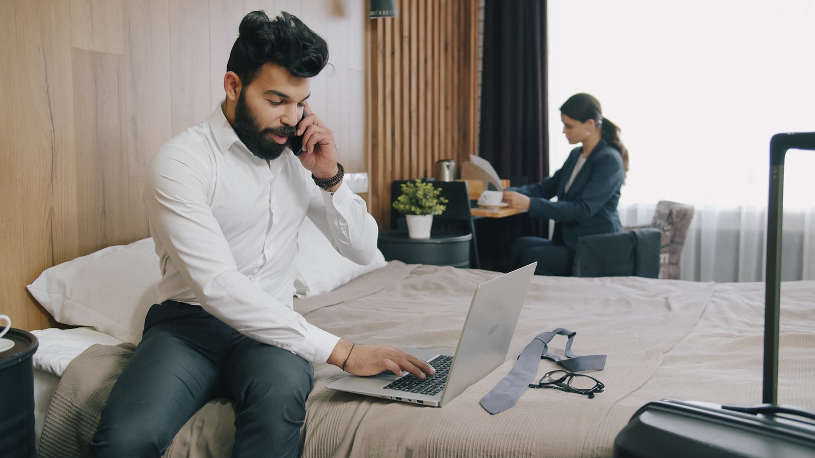 A man in a white shirt using a laptop while sitting on a hotel bed (photo: Vitaly Gariev / Unsplash)