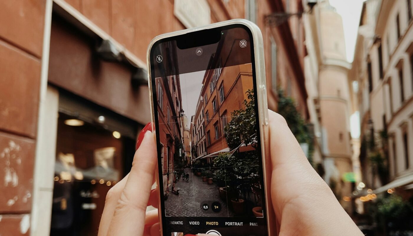 A hand holding a phone up to photograph a narrow European street lined with old buildings (photo: Valentyn Chernetskyi / Unsplash)