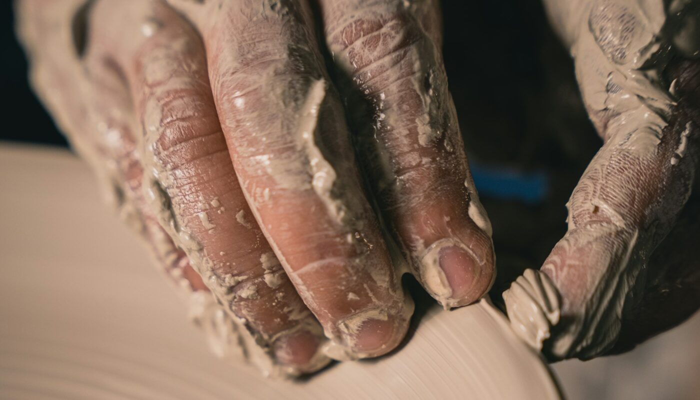 A potter’s clay-covered hands resting on a spinning wheel as a form takes shape (photo: Tuan Nguyen / Unsplash)