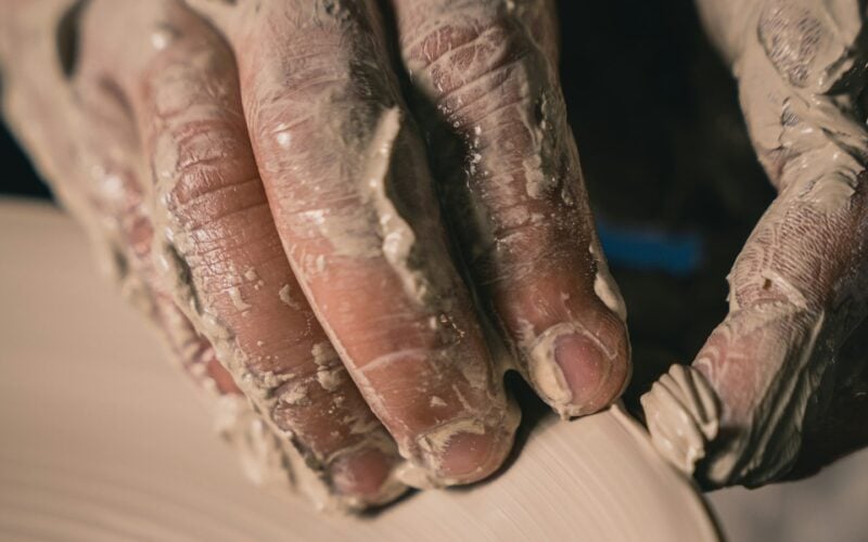 A potter’s clay-covered hands resting on a spinning wheel as a form takes shape (photo: Tuan Nguyen / Unsplash)