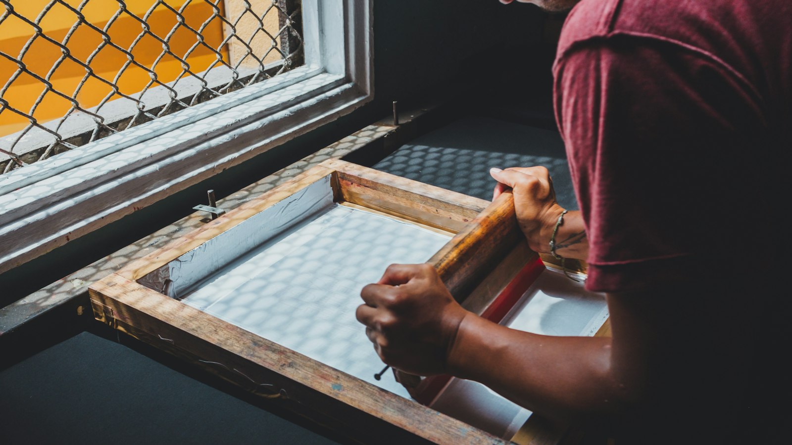 A printer lifting a screen-printing frame over a patterned sheet in a daylit workshop