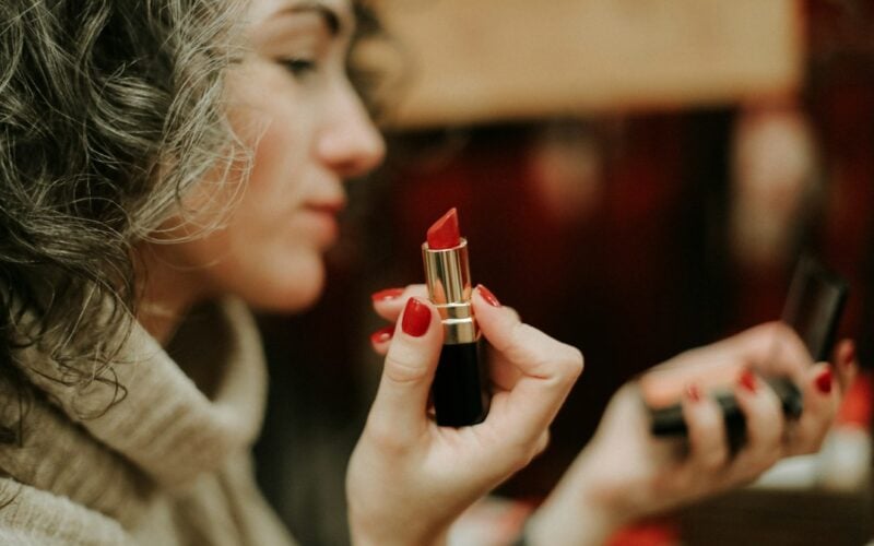 A woman holds up a red lipstick and a compact mirror in a warmly lit interior.