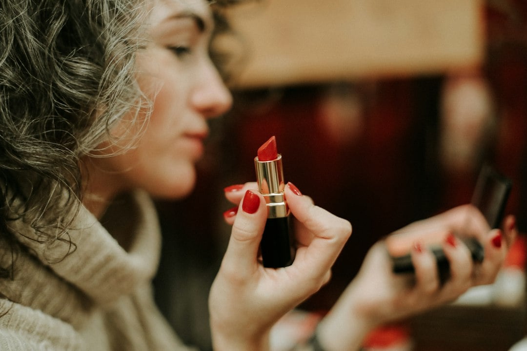 A woman holds up a red lipstick and a compact mirror in a warmly lit interior.