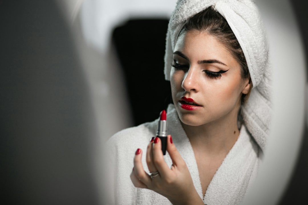 A woman in a bathrobe with a towel turban holds a red lipstick in front of a ring light.
