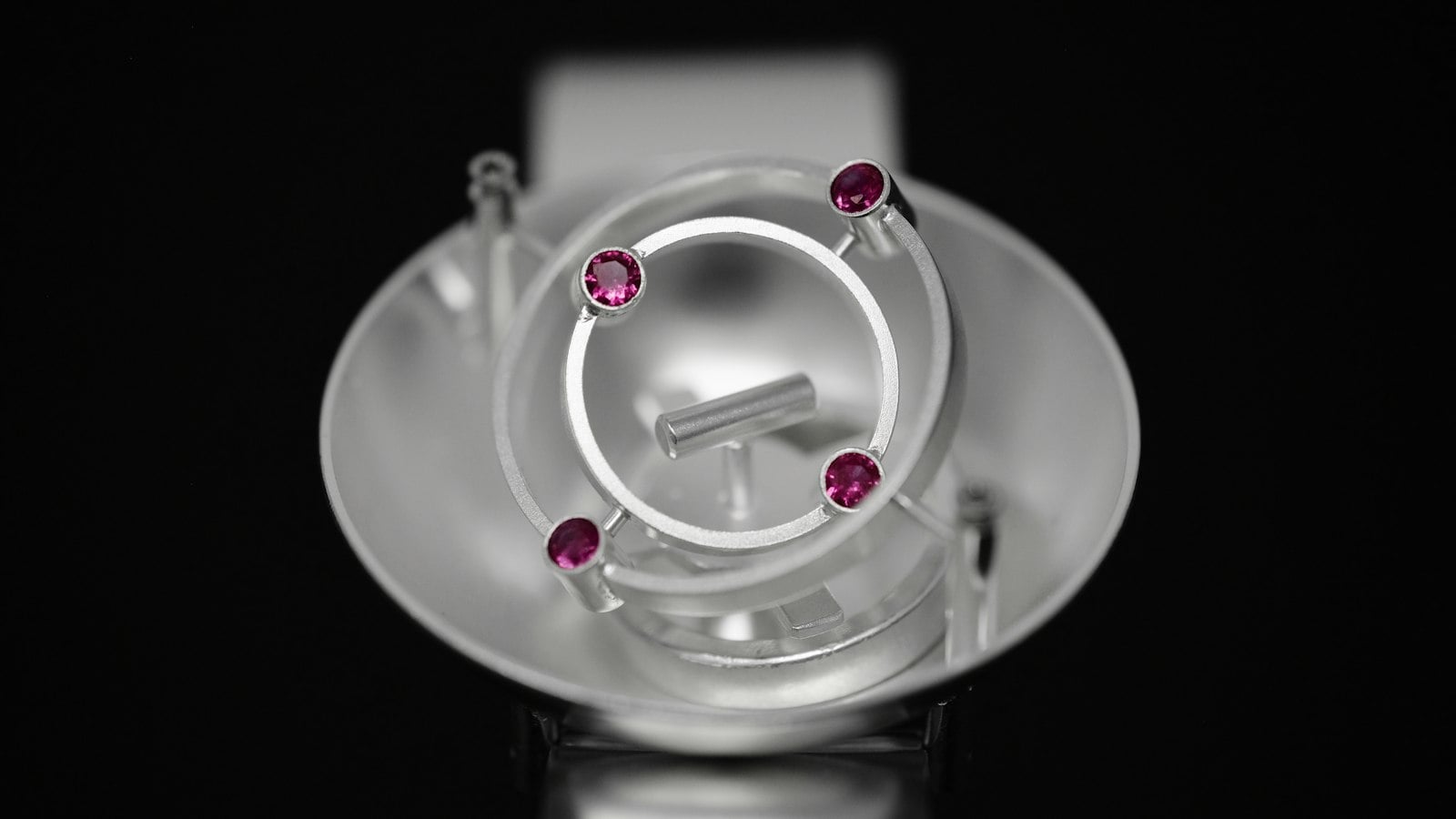 A silver ring on a display stand, set with four small pink stones, lit against a black background