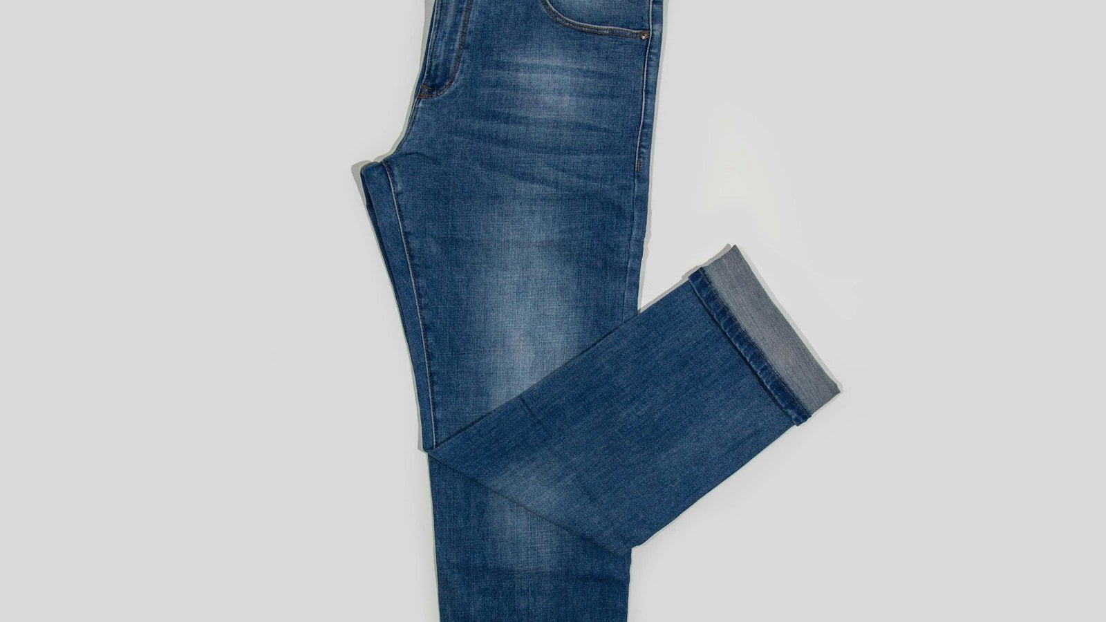 A single pair of mid-blue slim jeans laid flat with one leg folded and the hem cuffed (photo: TuanAnh Blue / Unsplash)