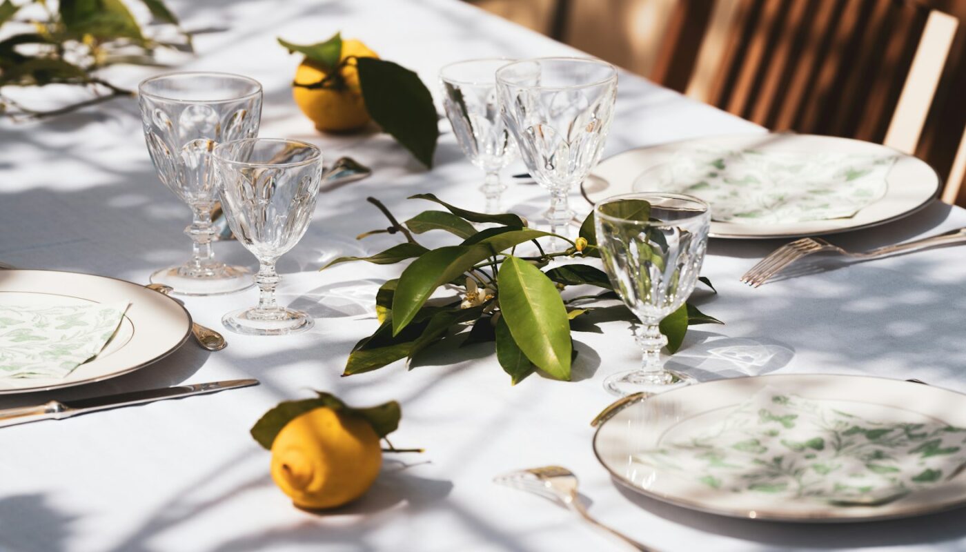 A white-clothed table set outdoors with cut-crystal glasses, patterned plates and sprigs of lemon leaves in dappled sunlight (photo: Guillaume de Germain / Unsplash)