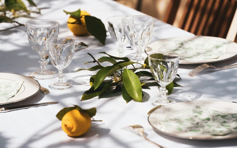A white-clothed table set outdoors with cut-crystal glasses, patterned plates and sprigs of lemon leaves in dappled sunlight (photo: Guillaume de Germain / Unsplash)