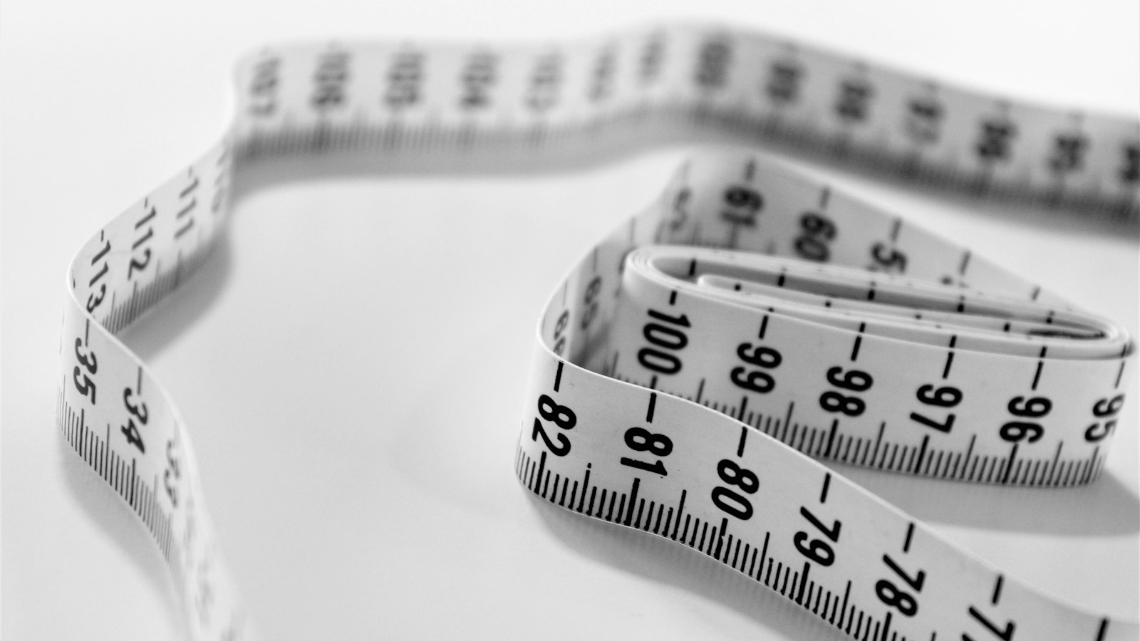 A close-up of a white tailor’s measuring tape curled on a pale surface (photo: Siora Photography / Unsplash)