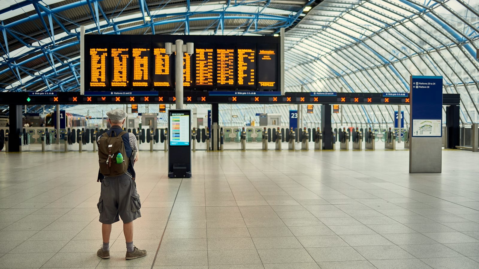 A traveller with a backpack standing in a bright, glass-roofed station hall looking up at the departure boards (photo: JJ Jordan / Unsplash)