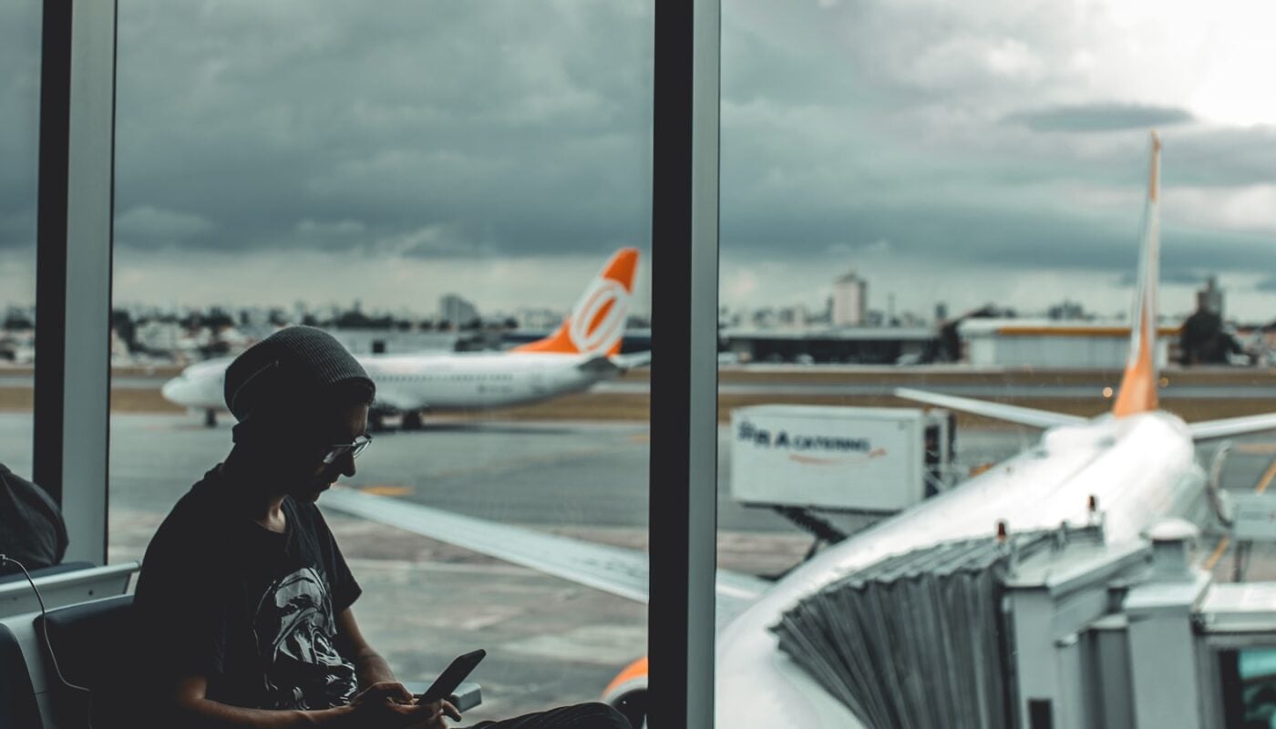 Traveller sitting at an airport gate looking at their phone, plane visible through the window (photo: Guilherme Stecanella / Unsplash)