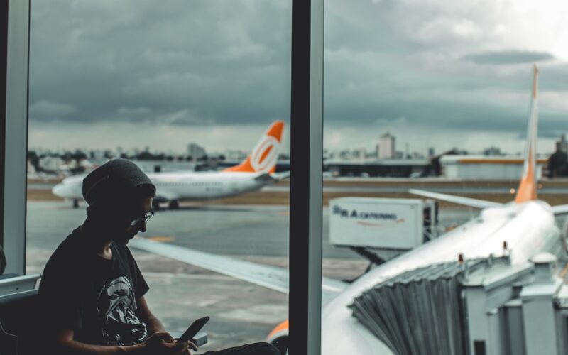 Traveller sitting at an airport gate looking at their phone, plane visible through the window (photo: Guilherme Stecanella / Unsplash)
