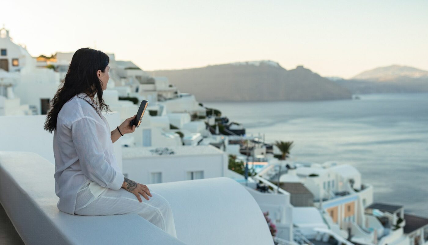 A traveller in white sitting on a whitewashed terrace and checking her phone above the sea and cliffs (photo: Anastase Maragos / Unsplash)
