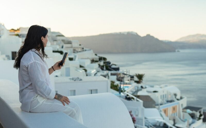 A traveller in white sitting on a whitewashed terrace and checking her phone above the sea and cliffs (photo: Anastase Maragos / Unsplash)