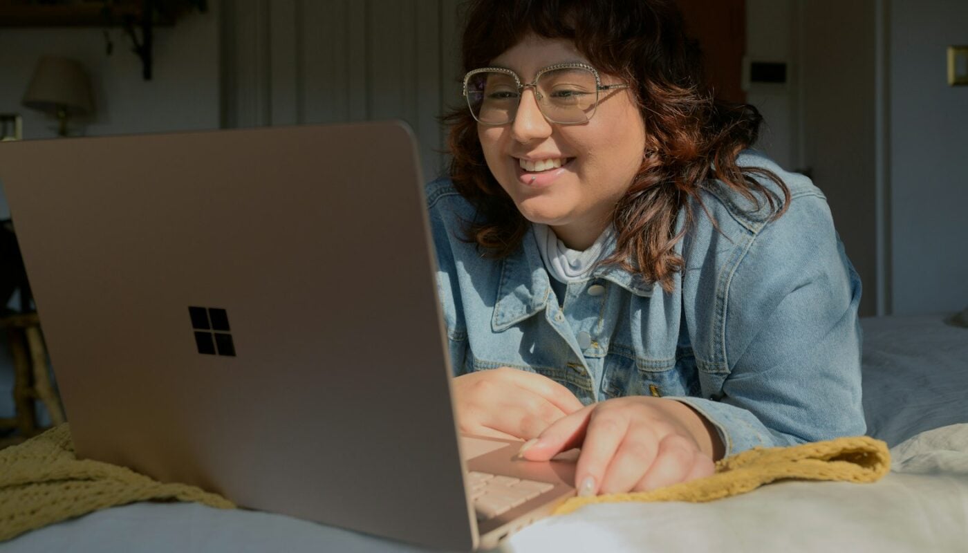 A woman in glasses leaning on a bed, smiling at a laptop screen in a dim room (photo: Surface / Unsplash)