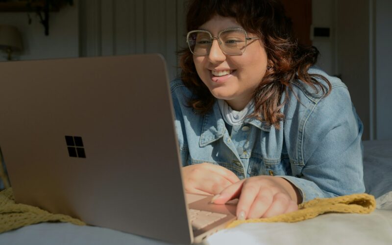 A woman in glasses leaning on a bed, smiling at a laptop screen in a dim room (photo: Surface / Unsplash)